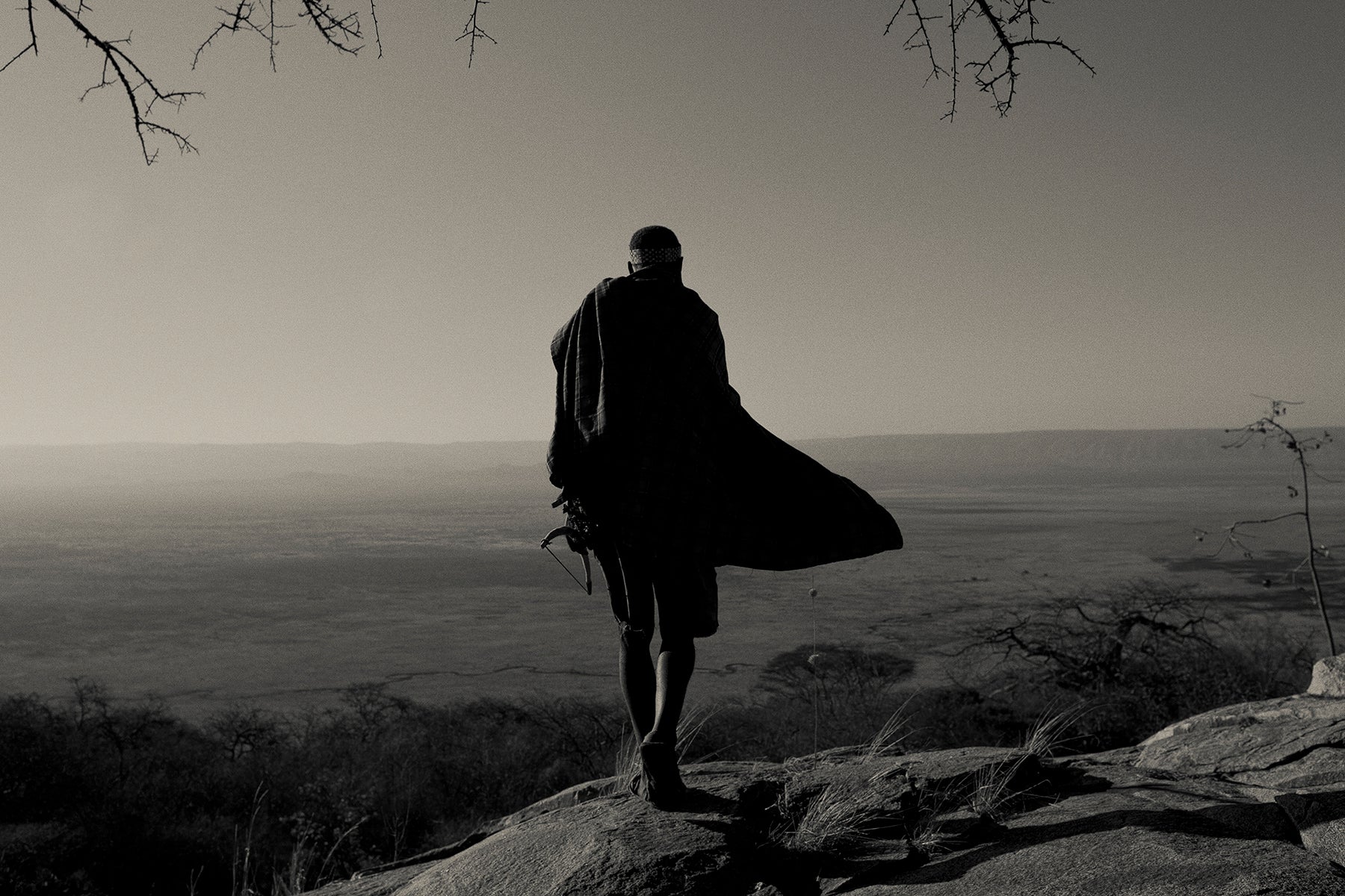 Elder standing on a rocky outcrop with a vast Tanzanian landscape and overcast sky.