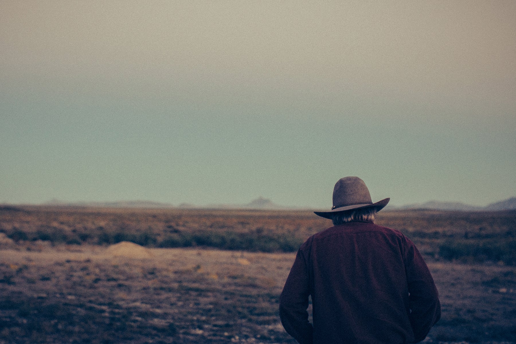 Person wearing a wide-brimmed hat standing in a vast, open Texas landscape with a muted color palette.